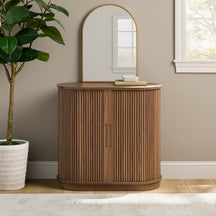 Wooden curve fluted accent cabinet w doors against a beige wall with a plant and arch mirror.