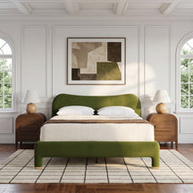 An olive green upholstered bed with a matching footboard and white bedding, styled in a bright, modern bedroom with white paneled walls, two wooden nightstands, table lamps, and a framed abstract painting above the headboard.