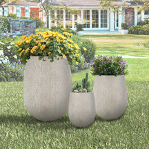 Three decorative planters with flowers and plants on a grassy lawn in front of a house.